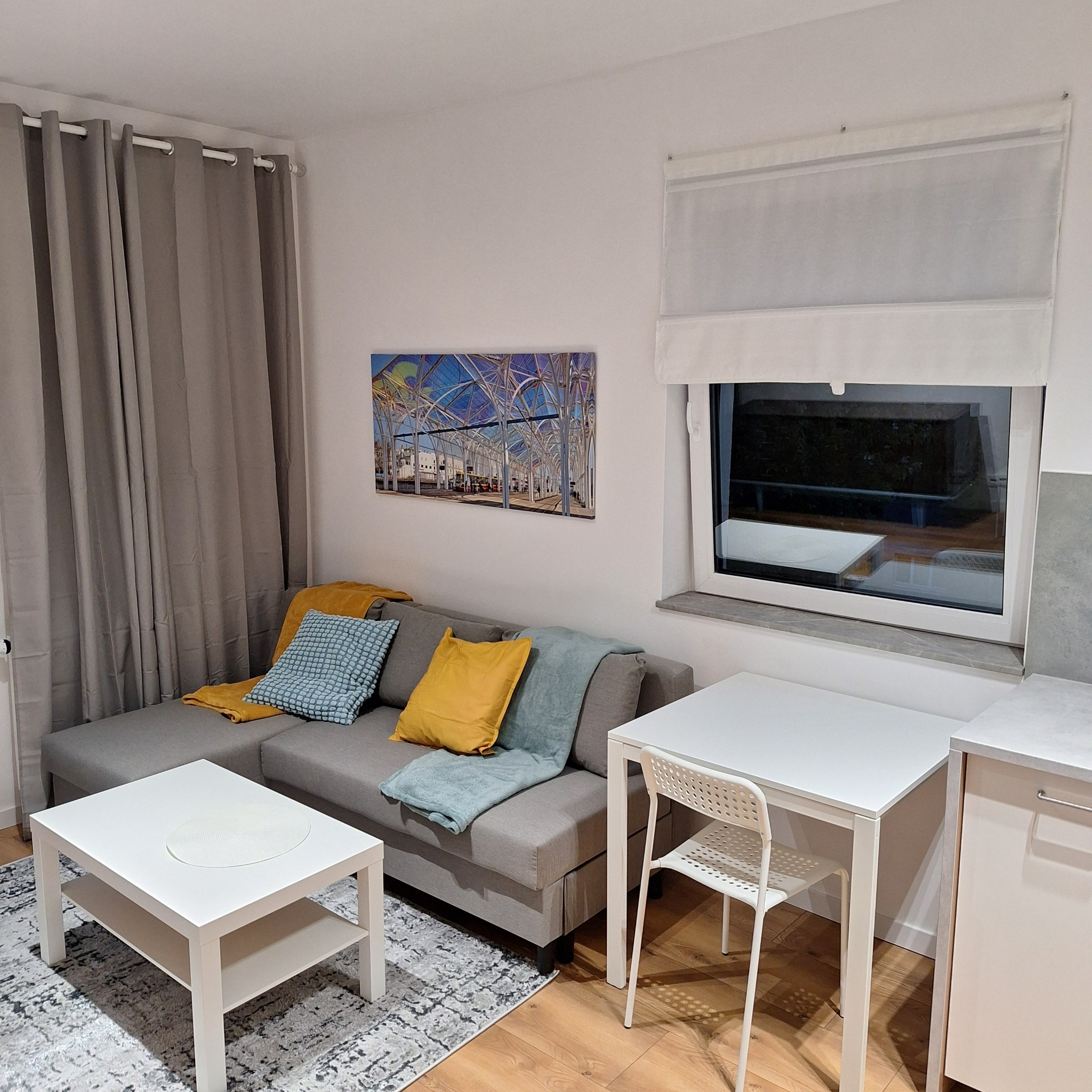 Jasne wnętrze mieszkania po home stagingu: szara sofa z poduszkami, biały stolik kawowy z okrągłą podkładką, biały stół i krzesło, obraz na ścianie, szare zasłony i drewniana podłoga.