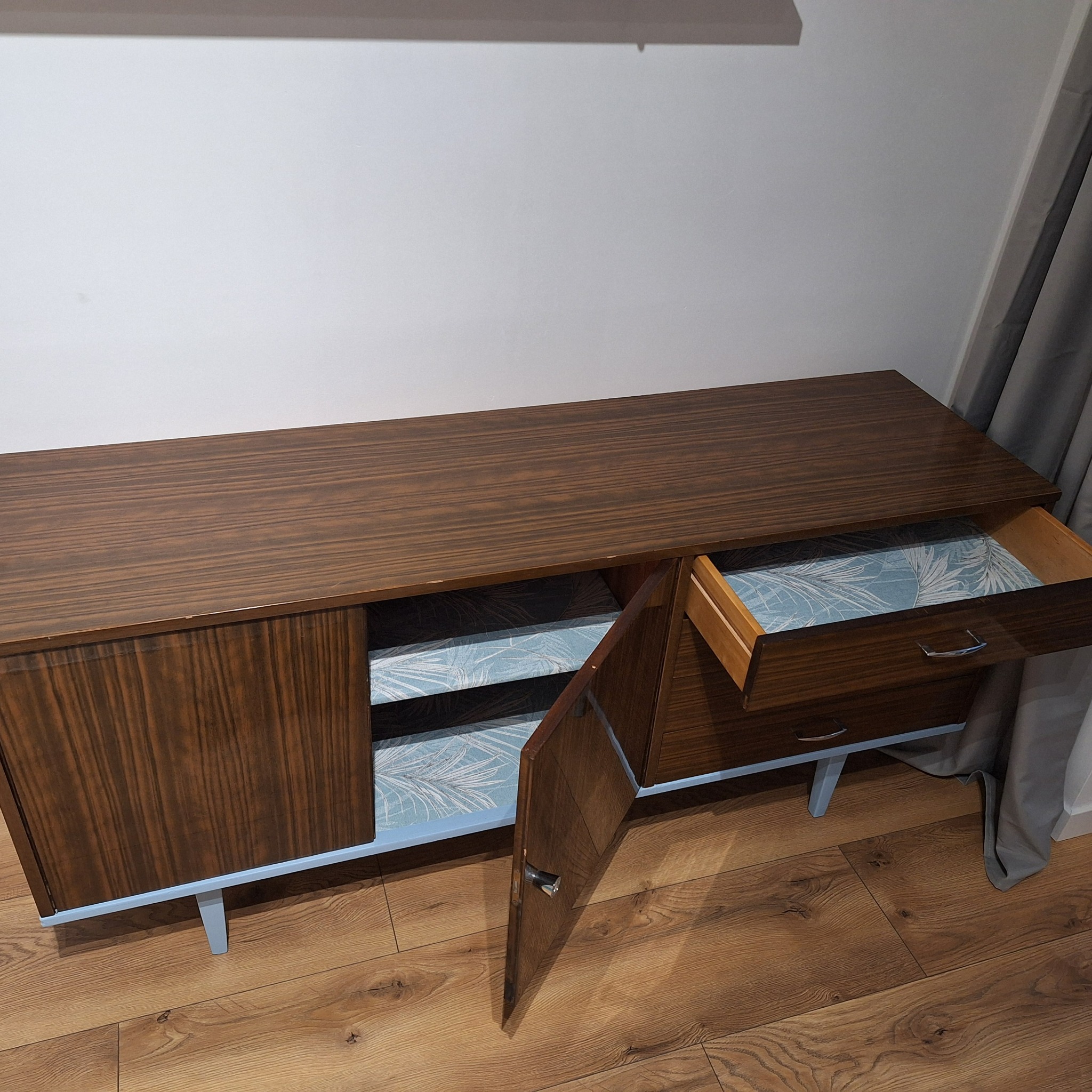 Odrestaurowana komoda w stylu vintage z otwartymi drzwiczkami i szufladą, wnętrze wyłożone tapetą w jasne liście, na drewnianej podłodze.