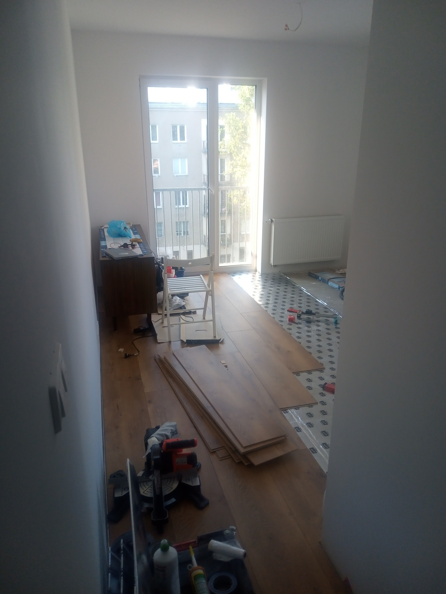 Układanie paneli podłogowych w mieszkaniu developerskim, widoczna piła ukośnica, folia izolacyjna i częściowo ułożone panele z widokiem na balkon.