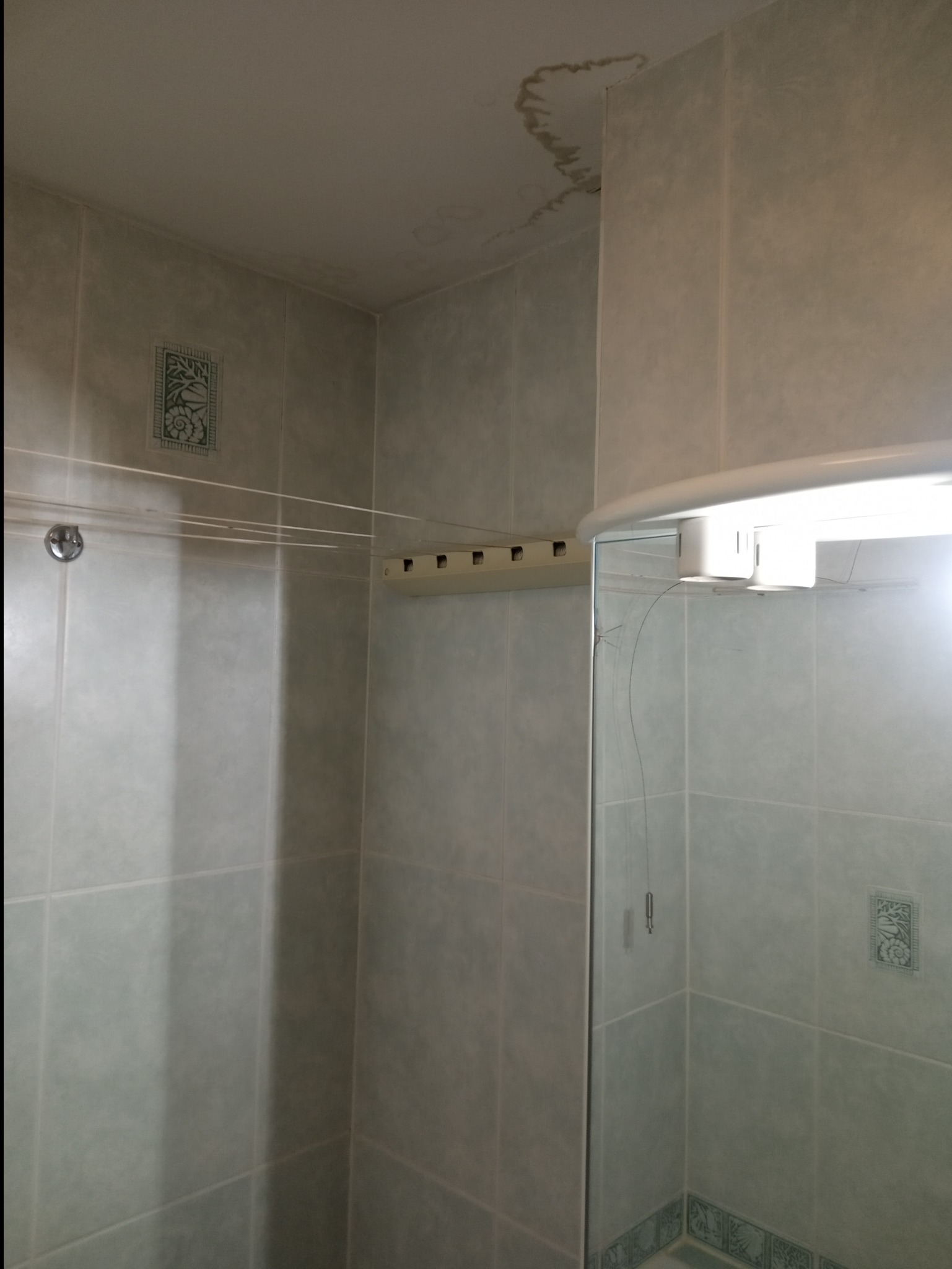 Łazienka z widocznymi plamami wilgoci na suficie, wiszącą suszarką sznurkową i kabiną prysznicową z pęknięciem na szkle.