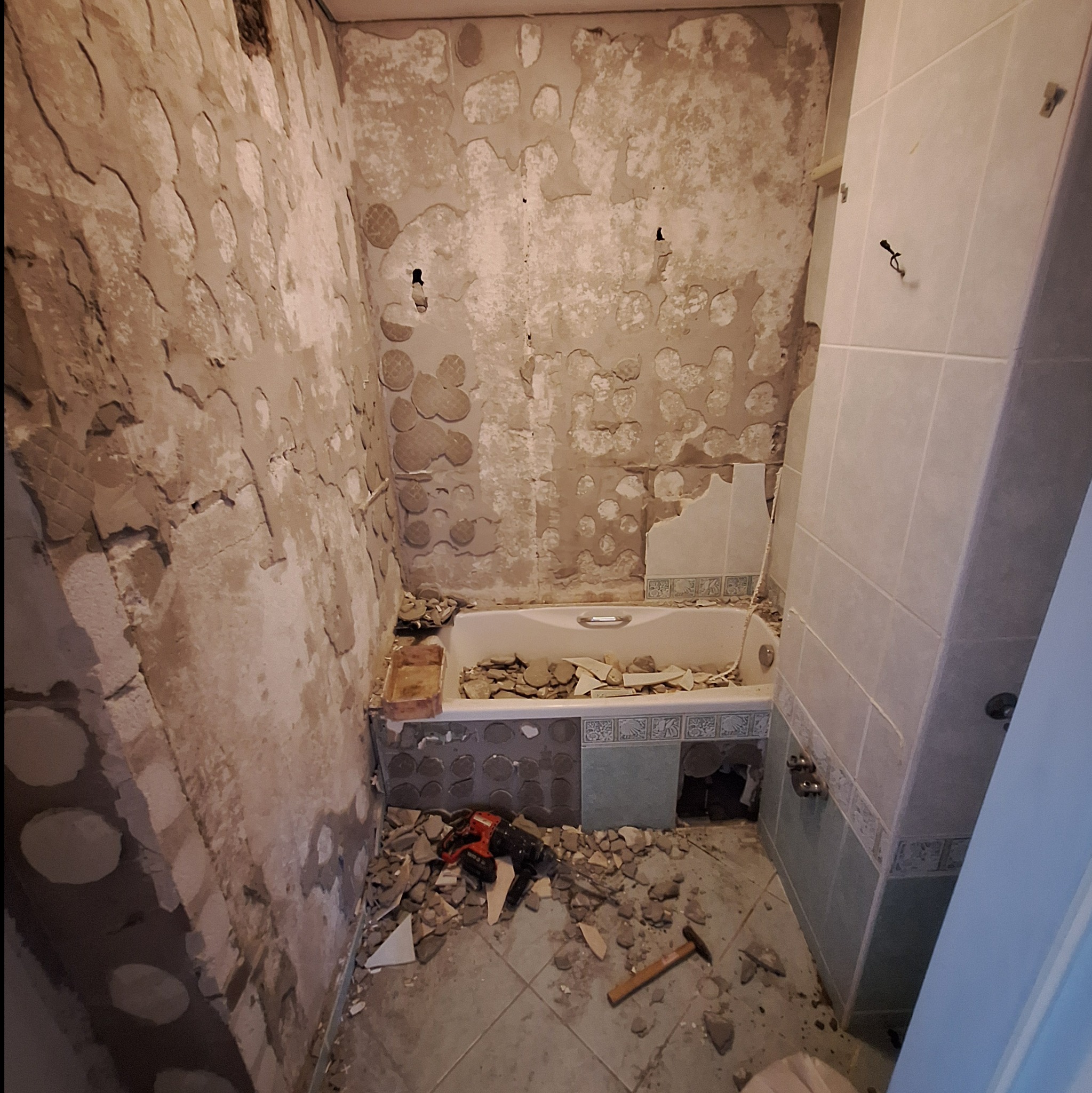 Demontaż płytek w łazience: widok na starą wannę z resztkami płytek, odspojone ściany z widocznymi warstwami podłoża, młotowiertarka i młotek na podłodze.