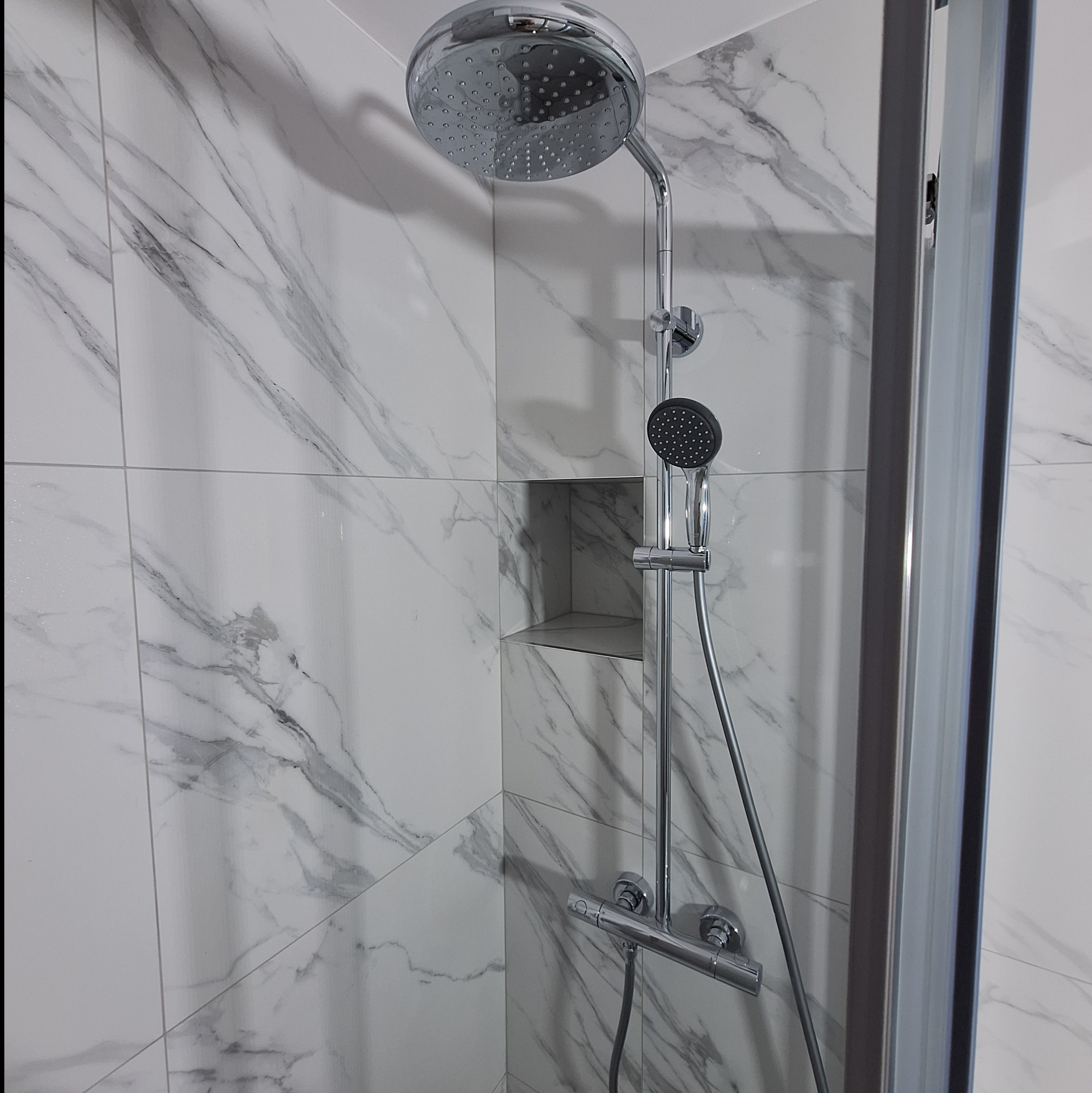 Nowoczesna kabina prysznicowa z chromowaną armaturą i deszczownicą na tle białych płytek imitujących marmur, z wnęką w ścianie.