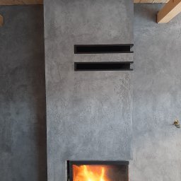 KAJGOR - Wykończony kominek z szarą, teksturowaną ścianą, drewnianą belką stropową i dwoma czarnymi, prostokątnymi wnękami dekoracyjnymi.