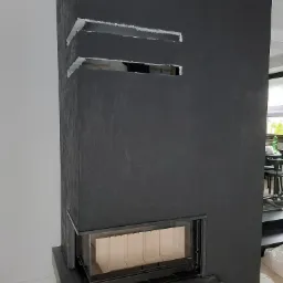 Ciemnoszary kominek z wkładem kominkowym, wykończony tynkiem strukturalnym, z widocznymi otworami wentylacyjnymi w górnej części, w trakcie prac wykończeniowych.