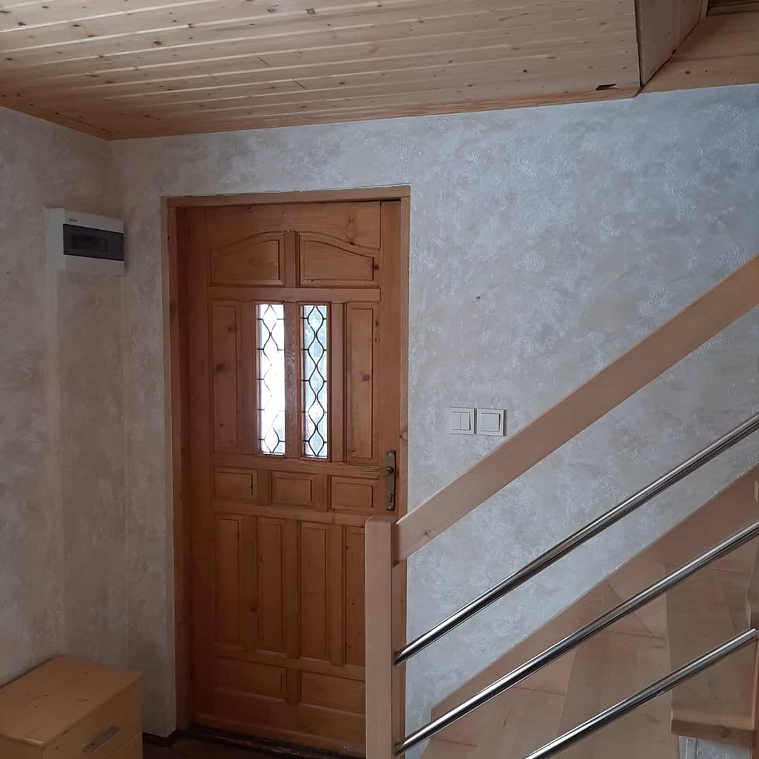 Wnętrze domu z drewnianą stolarką: drzwi z szybkami i schody z metalowymi balustradami, ściany wykończone dekoracyjnym tynkiem w odcieniach beżu i szarości, drewniany sufit.