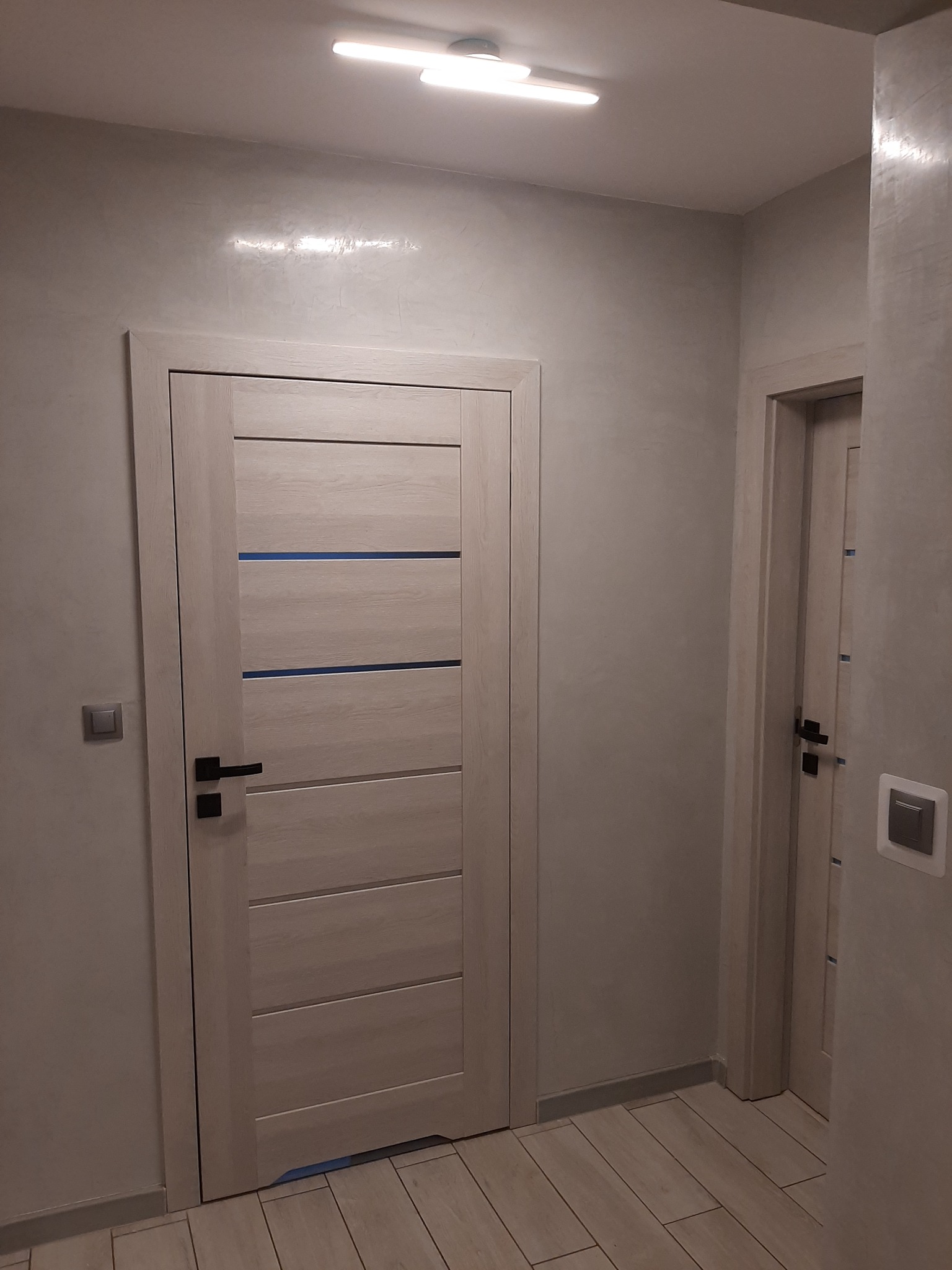 Ściana w odcieniach szarości z efektem dekoracyjnym, dwie drewniane drzwi z poziomymi, niebieskimi akcentami, podłoga wyłożona panelami w odcieniach beżu i szarości, oświetlenie sufitowe.