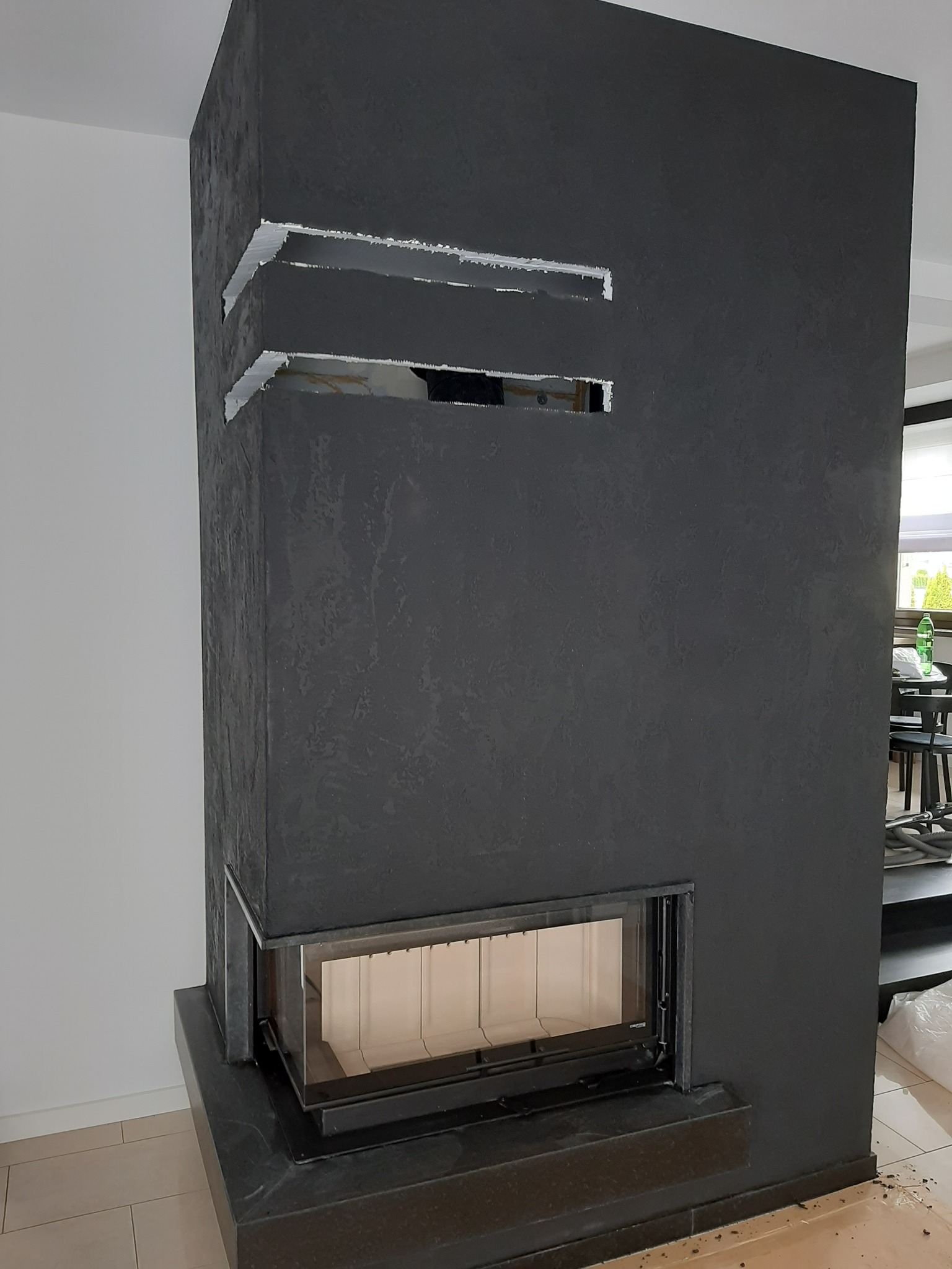 Ciemnoszary kominek z wkładem kominkowym, wykończony tynkiem strukturalnym, z widocznymi otworami wentylacyjnymi w górnej części, w trakcie prac wykończeniowych.