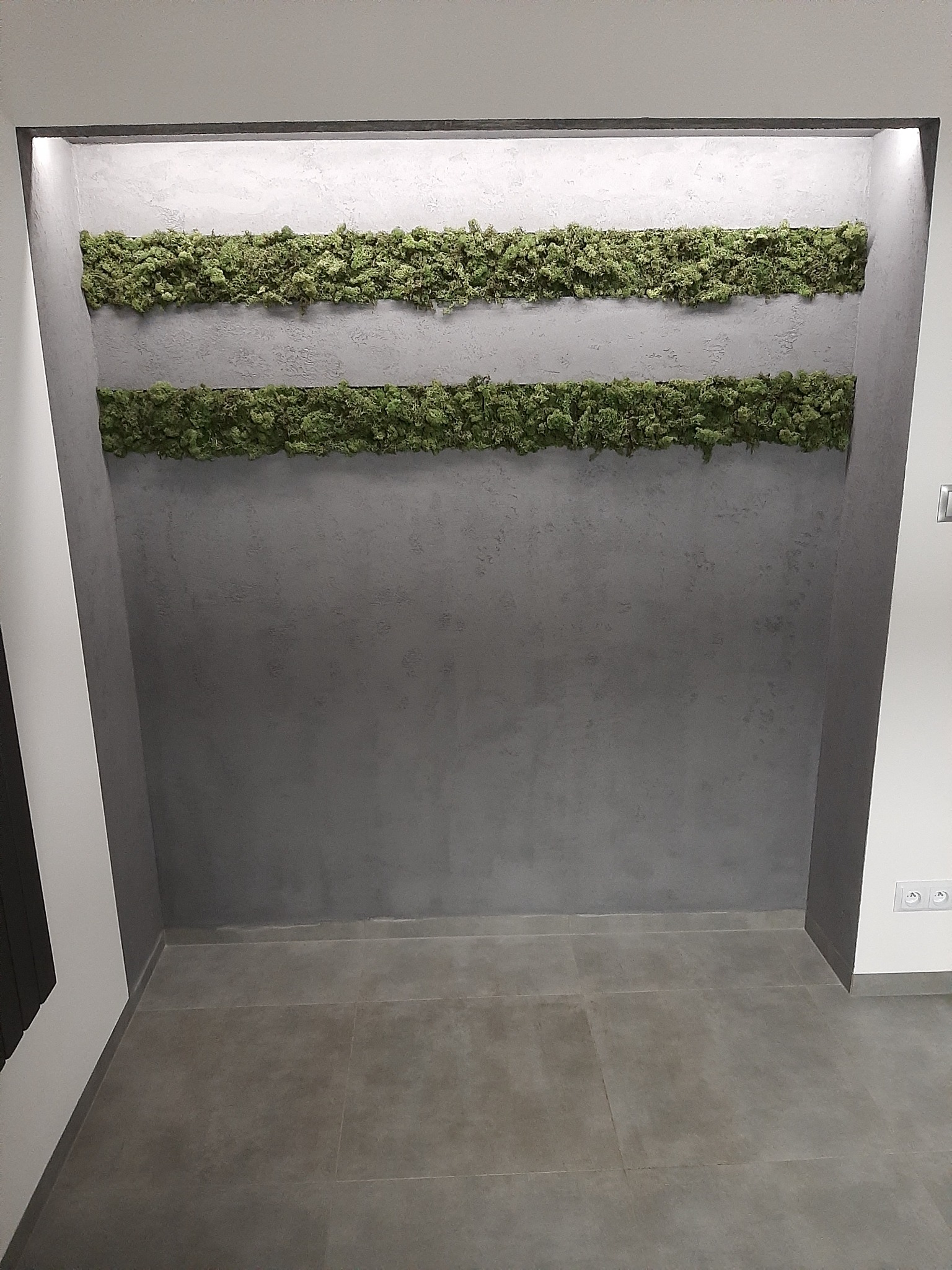 Szara ściana z dwoma poziomymi pasami zielonego mchu dekoracyjnego, oświetlona punktowo, z szarą podłogą z płytek.