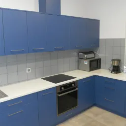 Niebieska zabudowa kuchenna z białym blatem, piekarnikiem i płytą indukcyjną, uzupełniona o mikrofalówkę i ekspres do kawy.