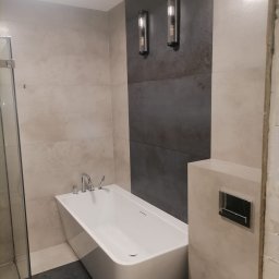 DAABAU Usługi Budowlane Remontowo - Wykończeniowe - Nowoczesna łazienka z białą wanną, szarymi płytkami na ścianie i ozdobnymi kinkietami, częściowo wykończona.