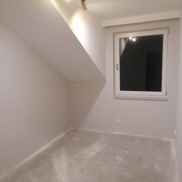 DAABAU Usługi Budowlane Remontowo - Wykończeniowe - Pomalowane na szaro poddasze ze skośnym sufitem, widoczne okno z białymi ramami i gniazdko elektryczne na ścianie, surowa podłoga przygotowana do dalszych prac wykończeniowych.