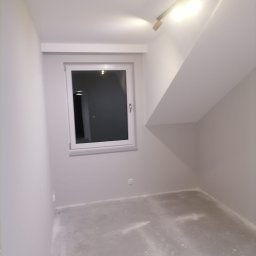 DAABAU Usługi Budowlane Remontowo - Wykończeniowe - Pomalowane na szaro wnętrze pokoju na poddaszu z oknem, widoczny skośny sufit i gładka, pomalowana podłoga.