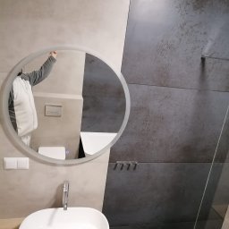 DAABAU Usługi Budowlane Remontowo - Wykończeniowe - Nowoczesna łazienka z umywalką nablatową, okrągłym lustrem z podświetleniem i szaro-beżowymi płytkami na ścianie, widoczne fragmenty armatury i toalety w odbiciu lustra.