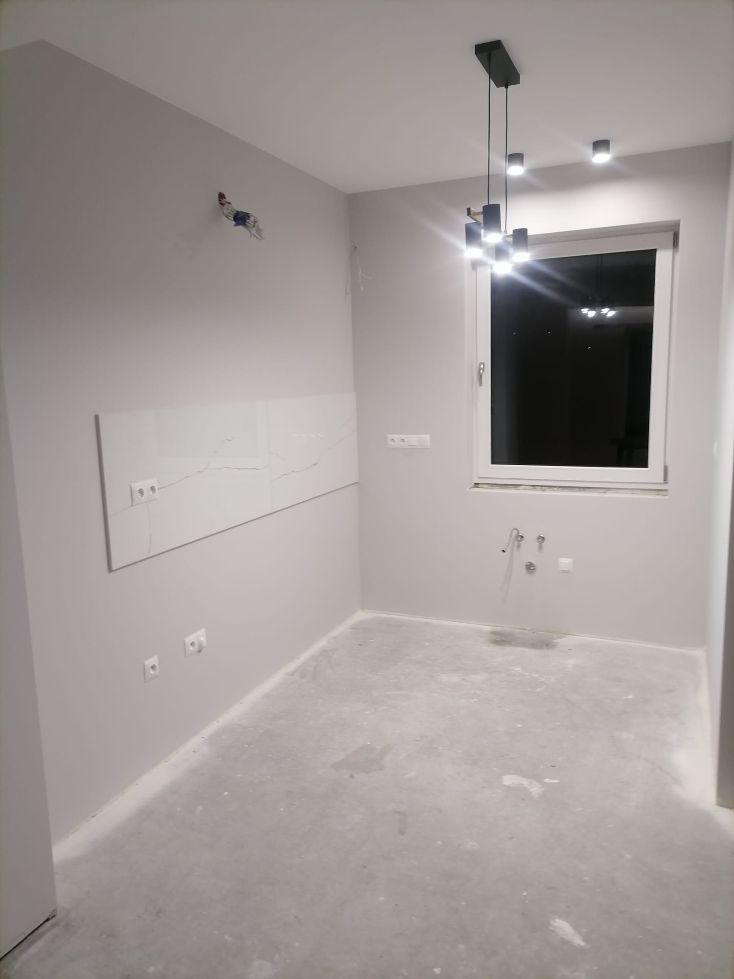 Wnętrze w trakcie prac: szare ściany, białe płytki imitujące marmur nad blatem, surowa podłoga, wisząca czarna lampa nad oknem.