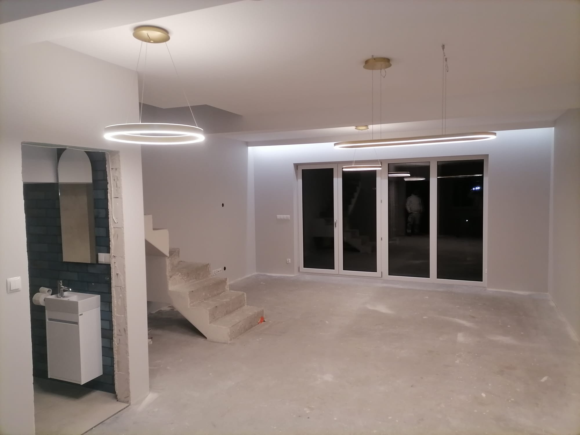 Surowy stan wnętrza z betonowymi schodami, trzema nowoczesnymi lampami wiszącymi i umywalką z niebieską mozaiką w tle.