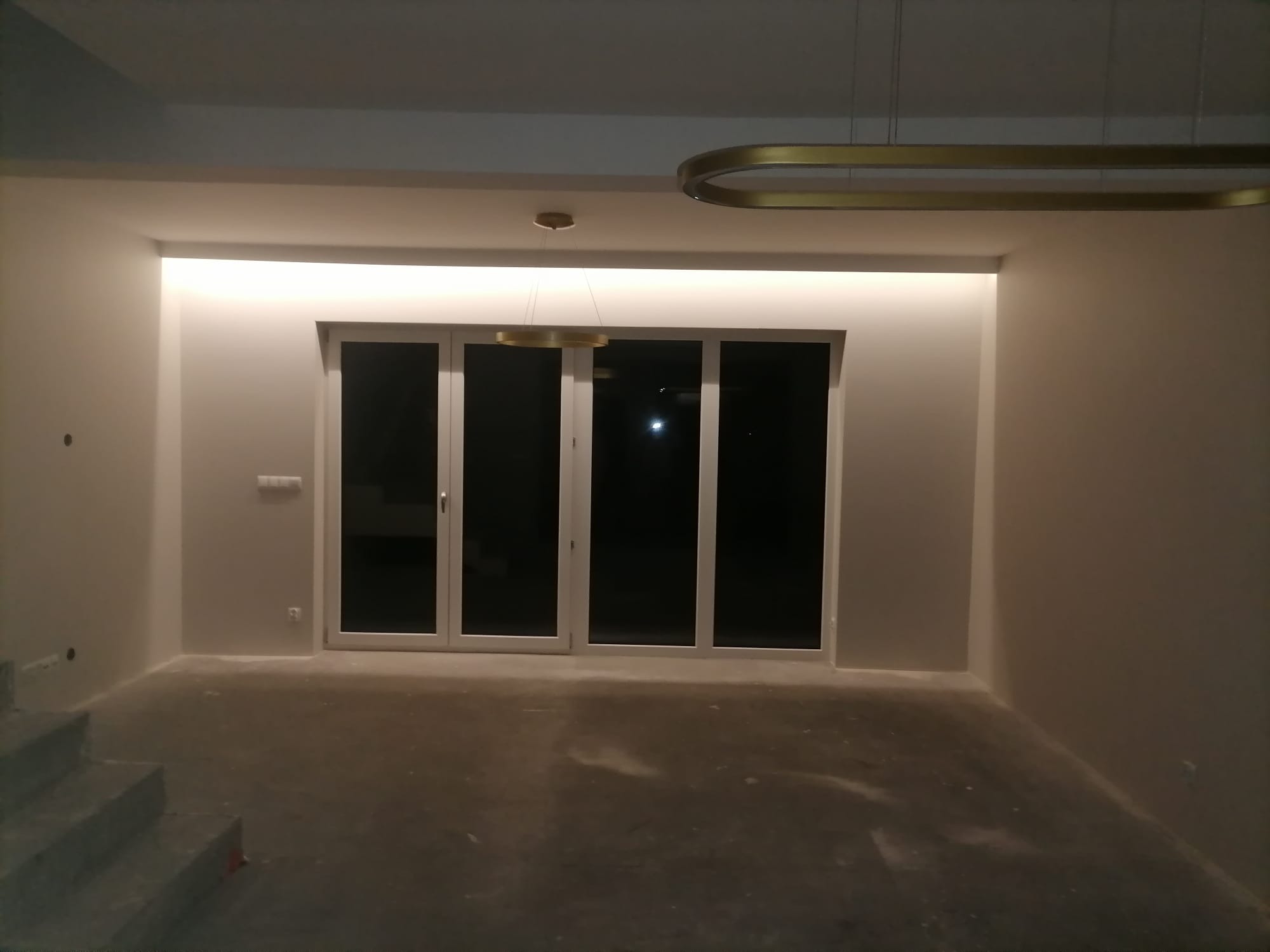 Wnętrze w trakcie prac wykończeniowych: gładkie, pomalowane ściany w odcieniach bieli, surowa podłoga, duże okno balkonowe z białymi ramami i nowoczesne, złote lampy sufitowe.