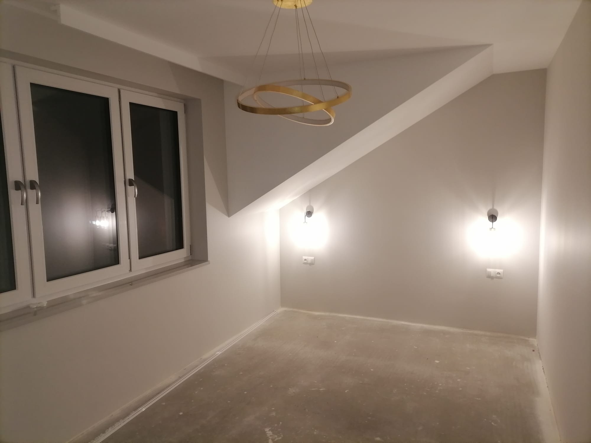 Pokój z szarymi ścianami, nowoczesną lampą pierścieniową i dwoma kinkietami; podłoga przygotowana do położenia nawierzchni.