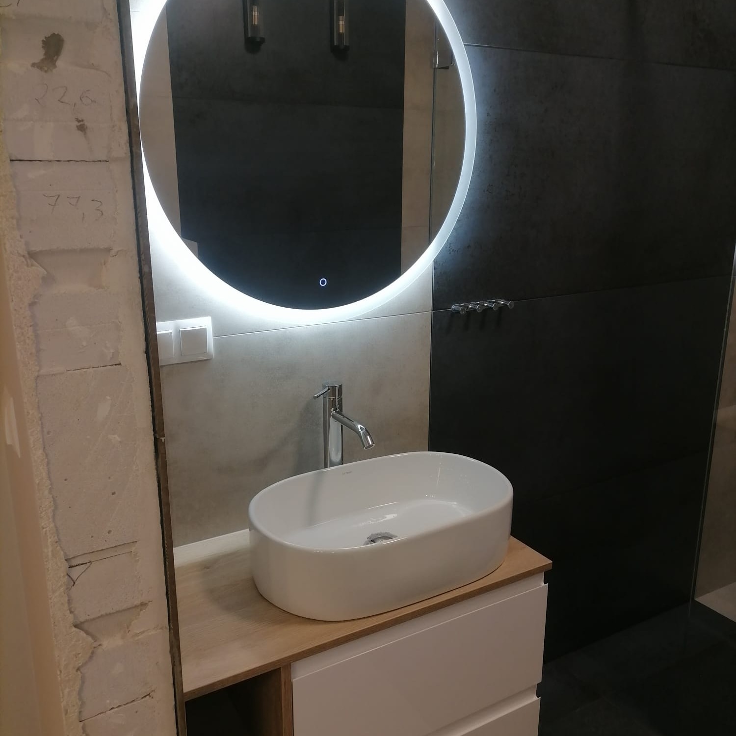 Łazienka w trakcie remontu: owalne umywalka nablatowa na szafce z blatem imitującym drewno, okrągłe lustro podświetlane LED, czarna ściana, surowa ściana z widocznymi numerami.