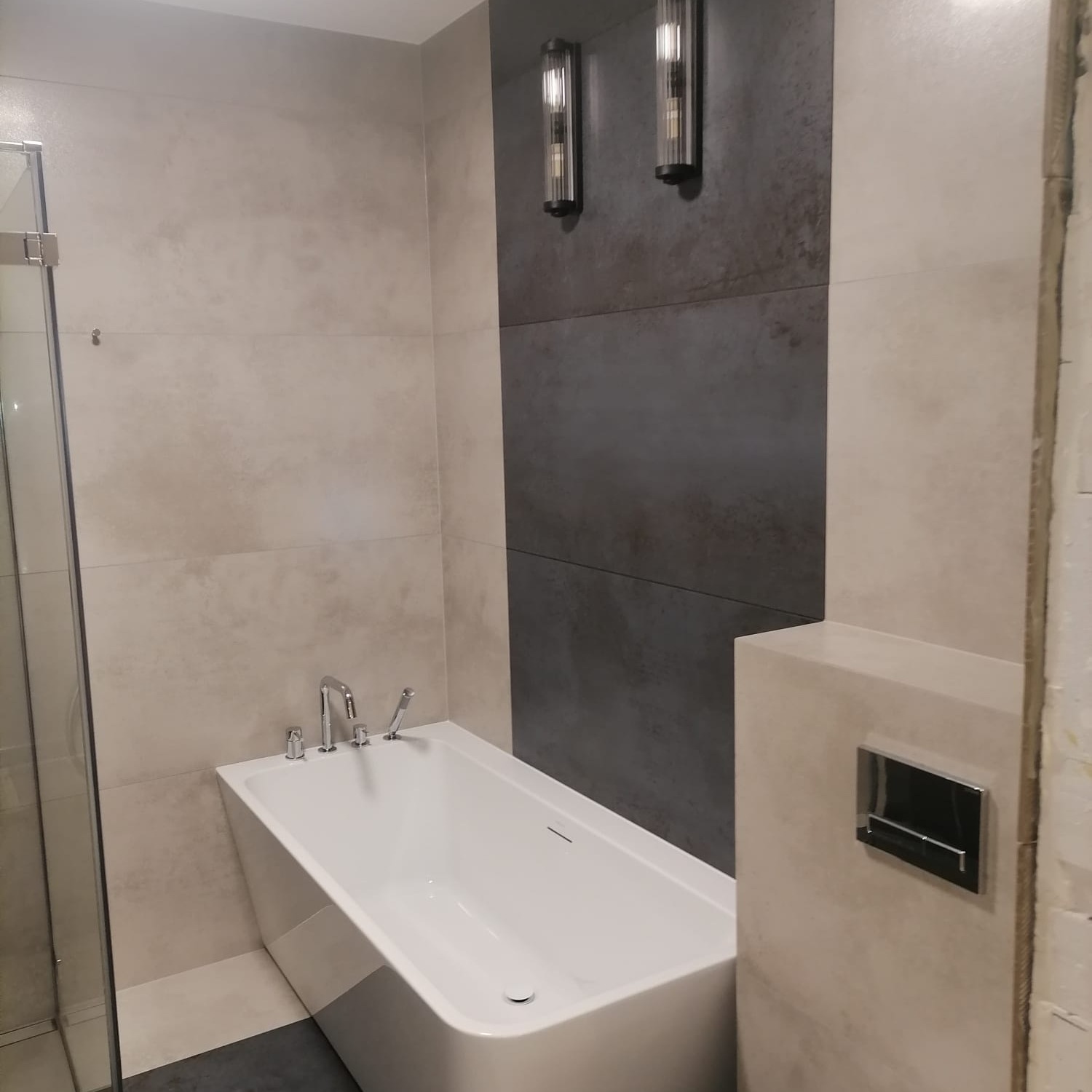 Nowoczesna łazienka z białą wanną, szarymi płytkami na ścianie i ozdobnymi kinkietami, częściowo wykończona.