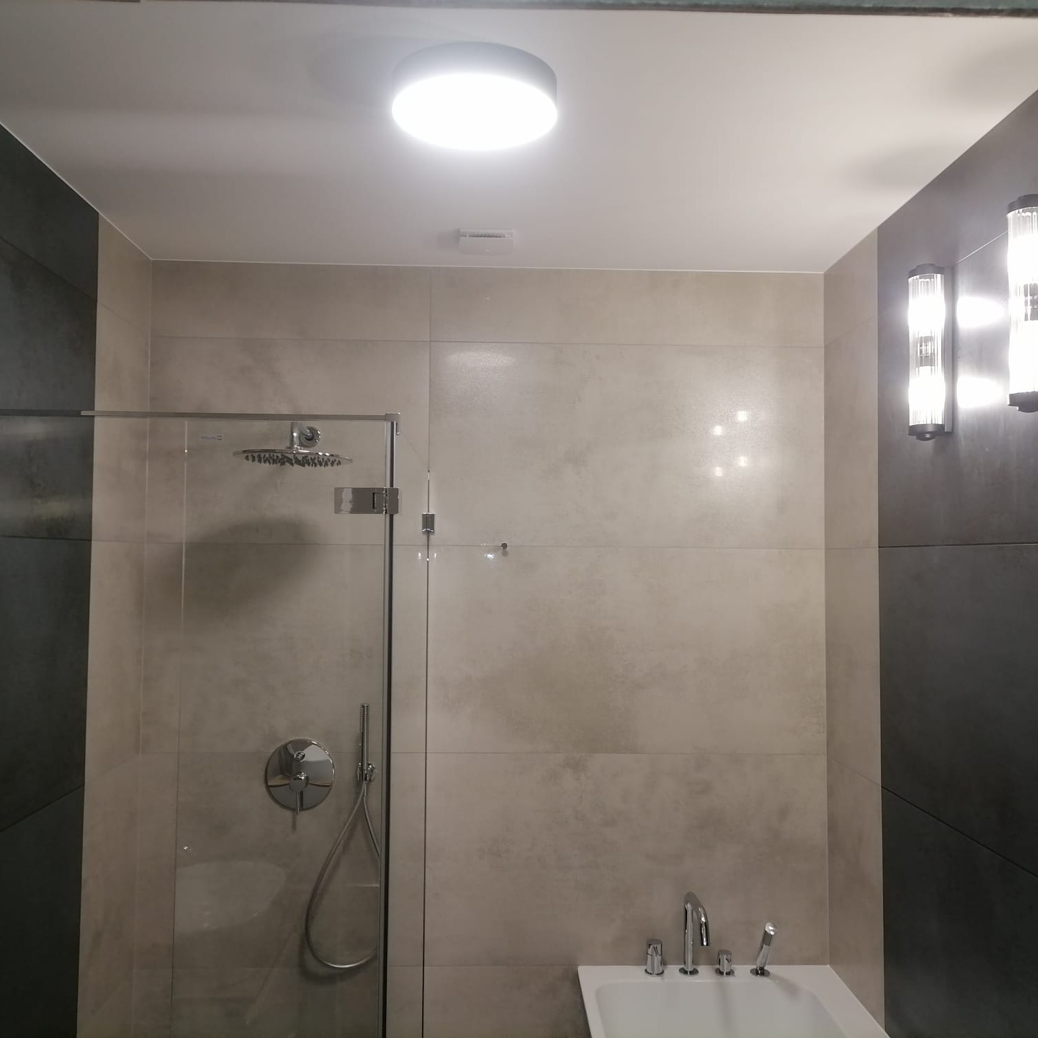 Nowoczesna łazienka z kabiną prysznicową typu walk-in, wykończona płytkami w odcieniach szarości i beżu, z chromowaną armaturą i oświetleniem punktowym.