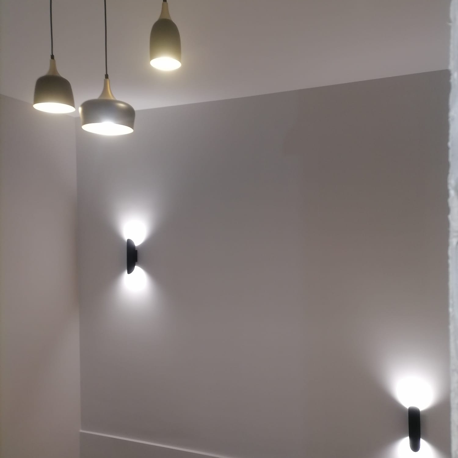 Nowoczesne oświetlenie wnętrza w Trzebnicy: trzy lampy wiszące w odcieniach szarości i złota oraz dwa kinkiety ścienne emitujące światło góra-dół na świeżo pomalowanej ścianie.
