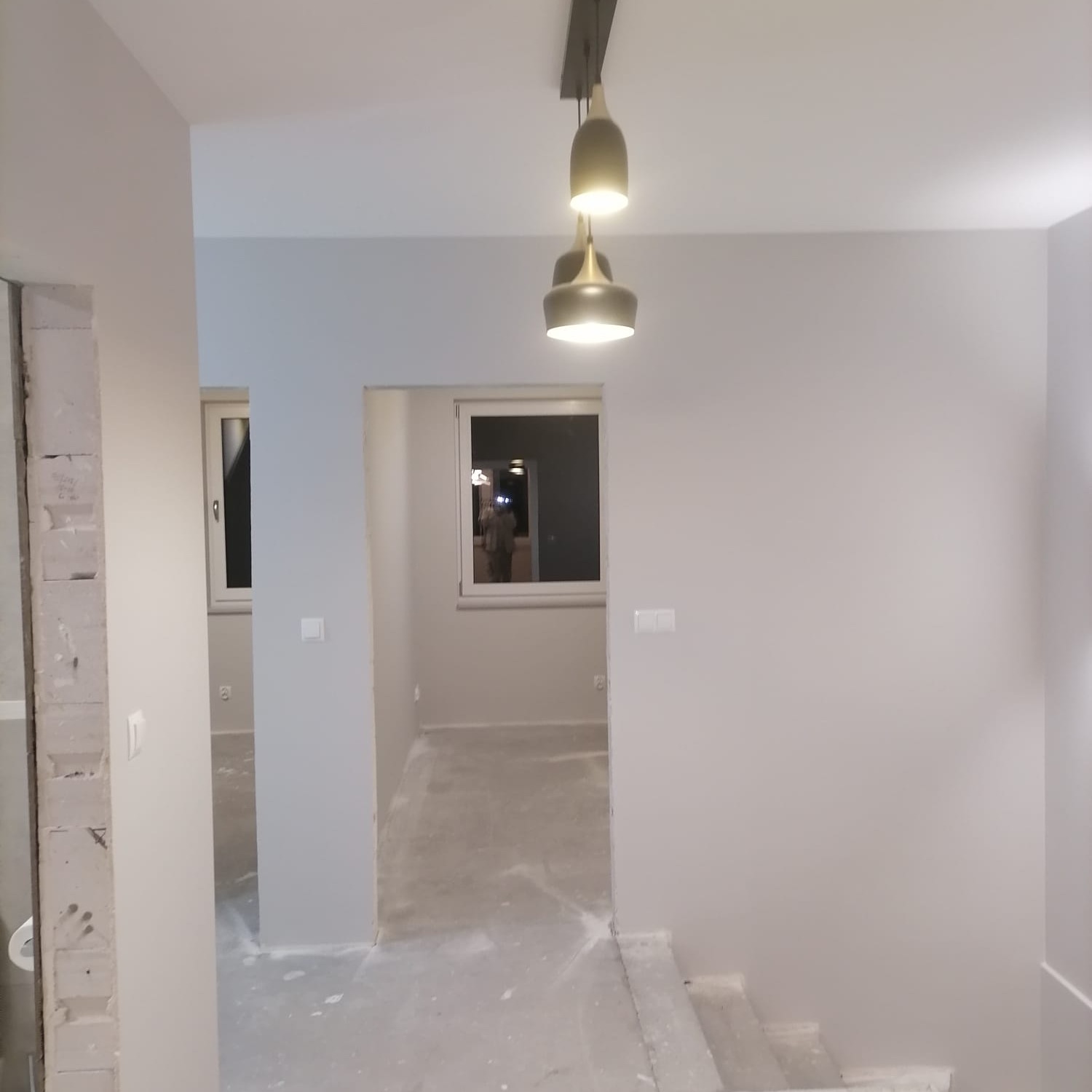 Wnętrze w trakcie prac wykończeniowych: świeżo pomalowane ściany w odcieniach szarości, surowa podłoga, nowoczesna lampa wisząca i widoczne okna w tle.