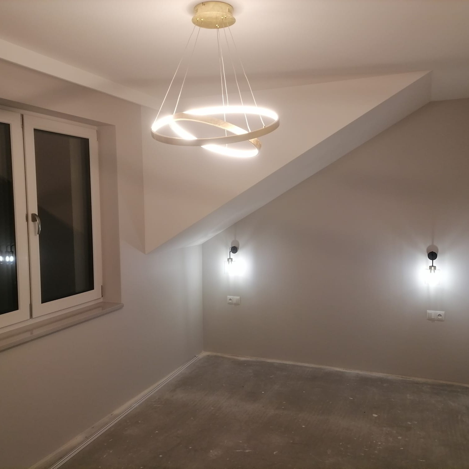 Pokój ze skośnym sufitem, oświetlony nowoczesną lampą pierścieniową i dwoma kinkietami, ściany pomalowane na szaro, surowa podłoga przed wykończeniem.