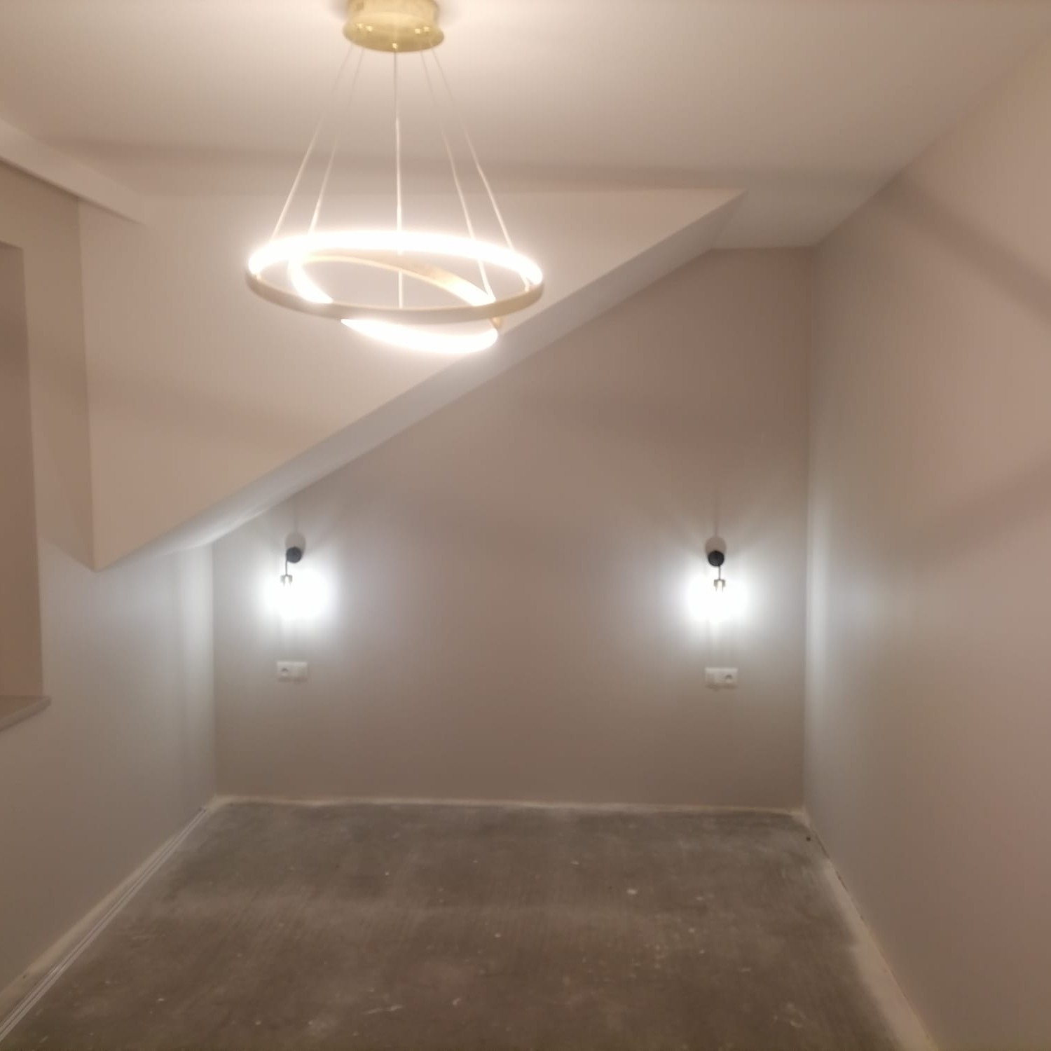 Poddasze po remoncie z gładkimi, pomalowanymi na szaro ścianami, nowoczesną, okrągłą lampą wiszącą i dwoma kinkietami po bokach. Podłoga przygotowana do dalszych prac wykończeniowych.