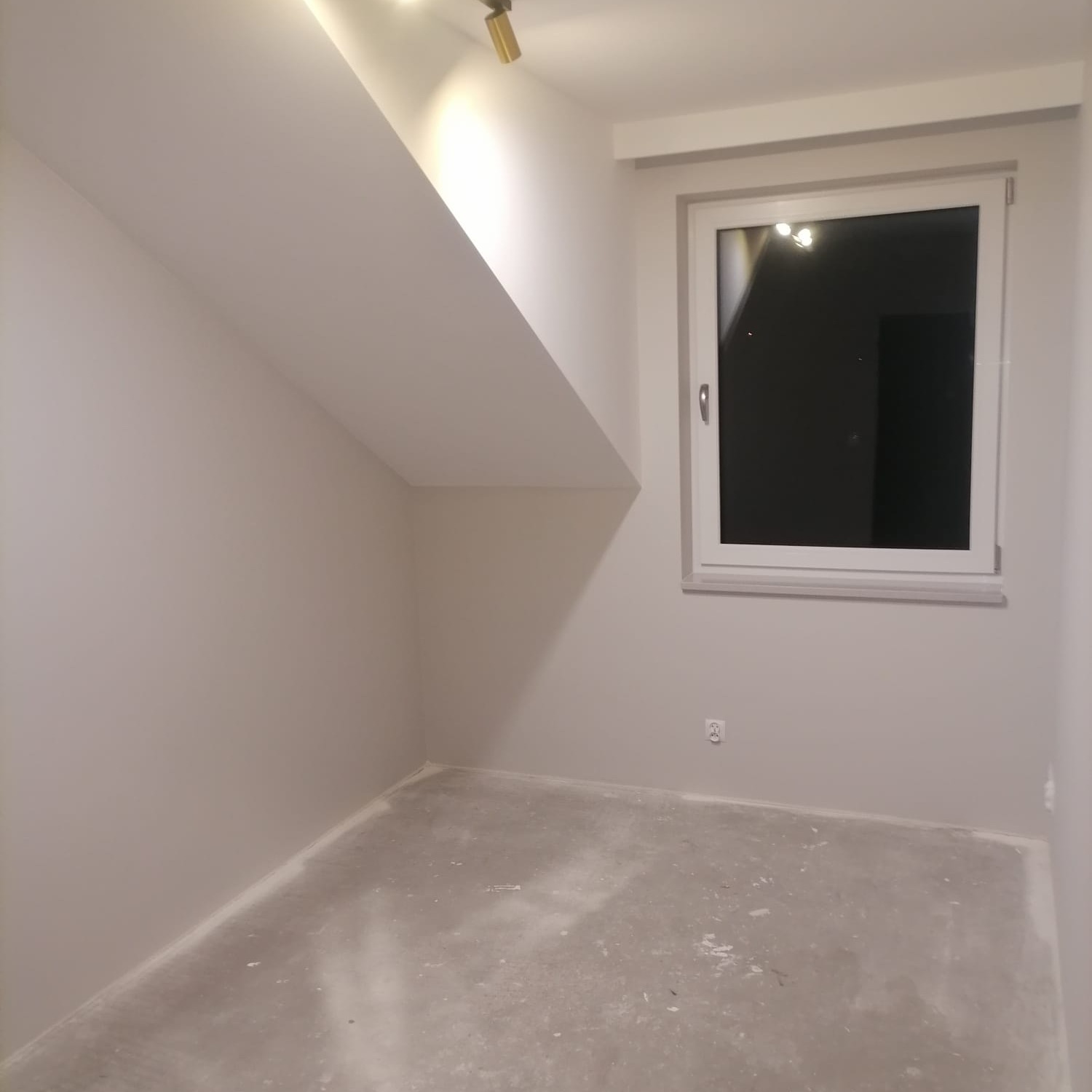 Pomalowane na szaro poddasze ze skośnym sufitem, widoczne okno z białymi ramami i gniazdko elektryczne na ścianie, surowa podłoga przygotowana do dalszych prac wykończeniowych.