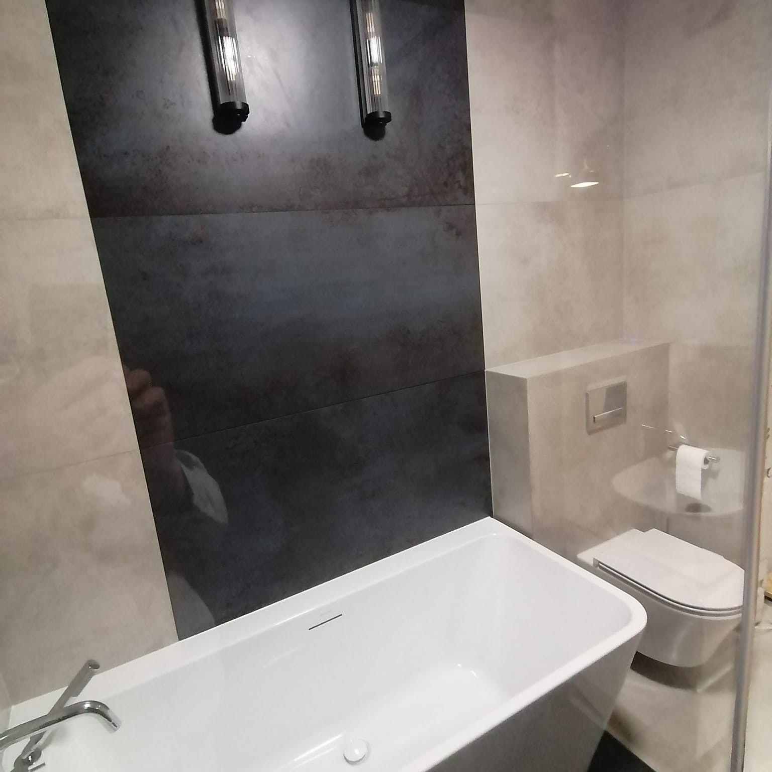 Nowoczesna łazienka z wanną wolnostojącą, toaletą podwieszaną i ścianą z ciemnoszarych płytek podświetloną pionowymi lampami.