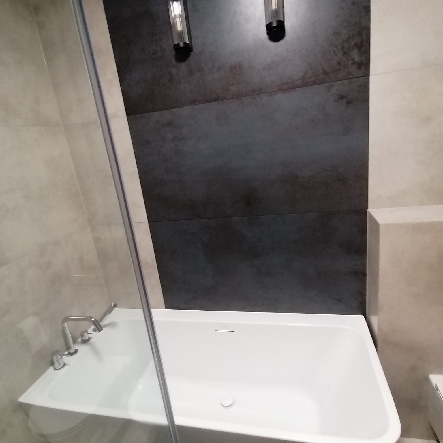 Nowoczesna biała wanna z chromowaną armaturą i szklanym parawanem, na tle ściany wyłożonej płytkami w odcieniach szarości i beżu oraz dwóch podłużnych lamp ściennych.