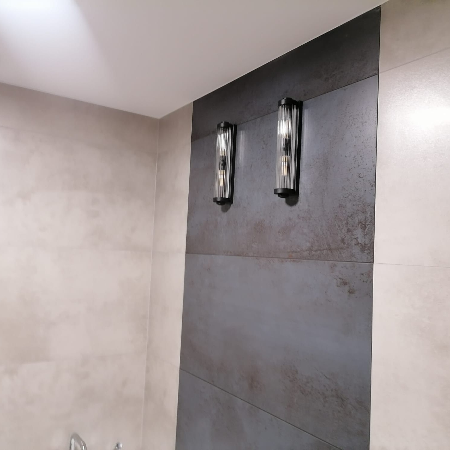 Dwie szklane lampy ścienne z czarnymi elementami, zamontowane na ścianie z płytek w odcieniach szarości i beżu, w łazience.