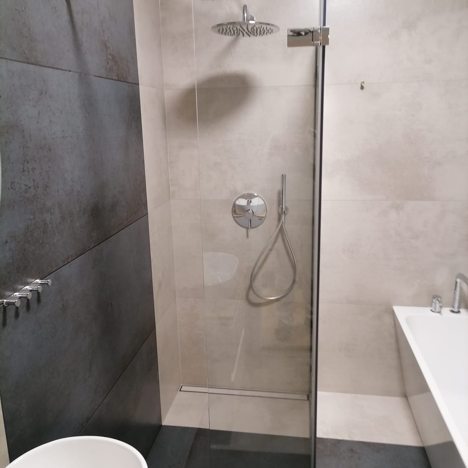 Nowoczesna łazienka z kabiną prysznicową typu walk-in, szare płytki na ścianie, deszczownica i bateria podtynkowa, widoczny fragment białej wanny.