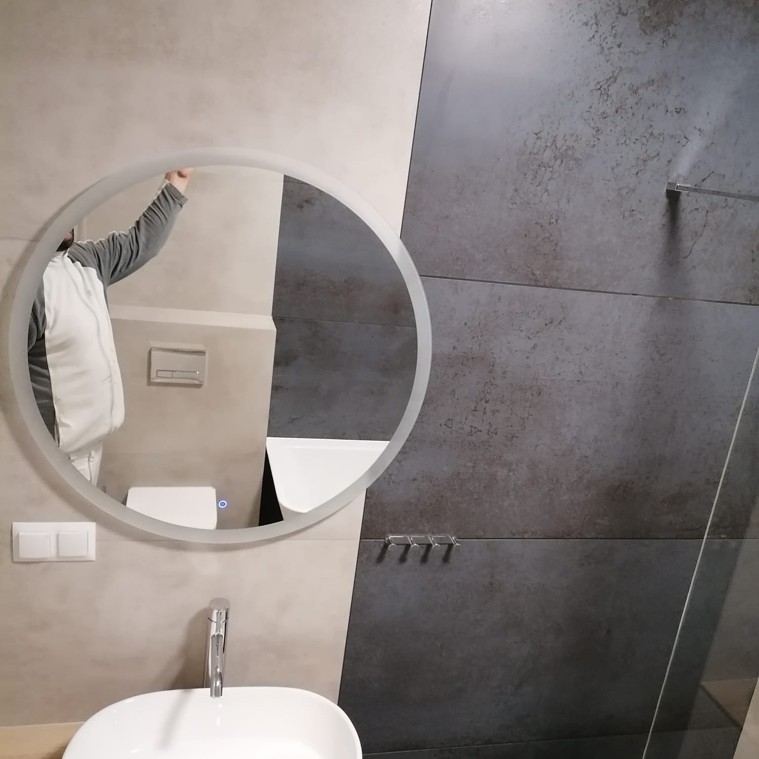 Nowoczesna łazienka z umywalką nablatową, okrągłym lustrem z podświetleniem i szaro-beżowymi płytkami na ścianie, widoczne fragmenty armatury i toalety w odbiciu lustra.