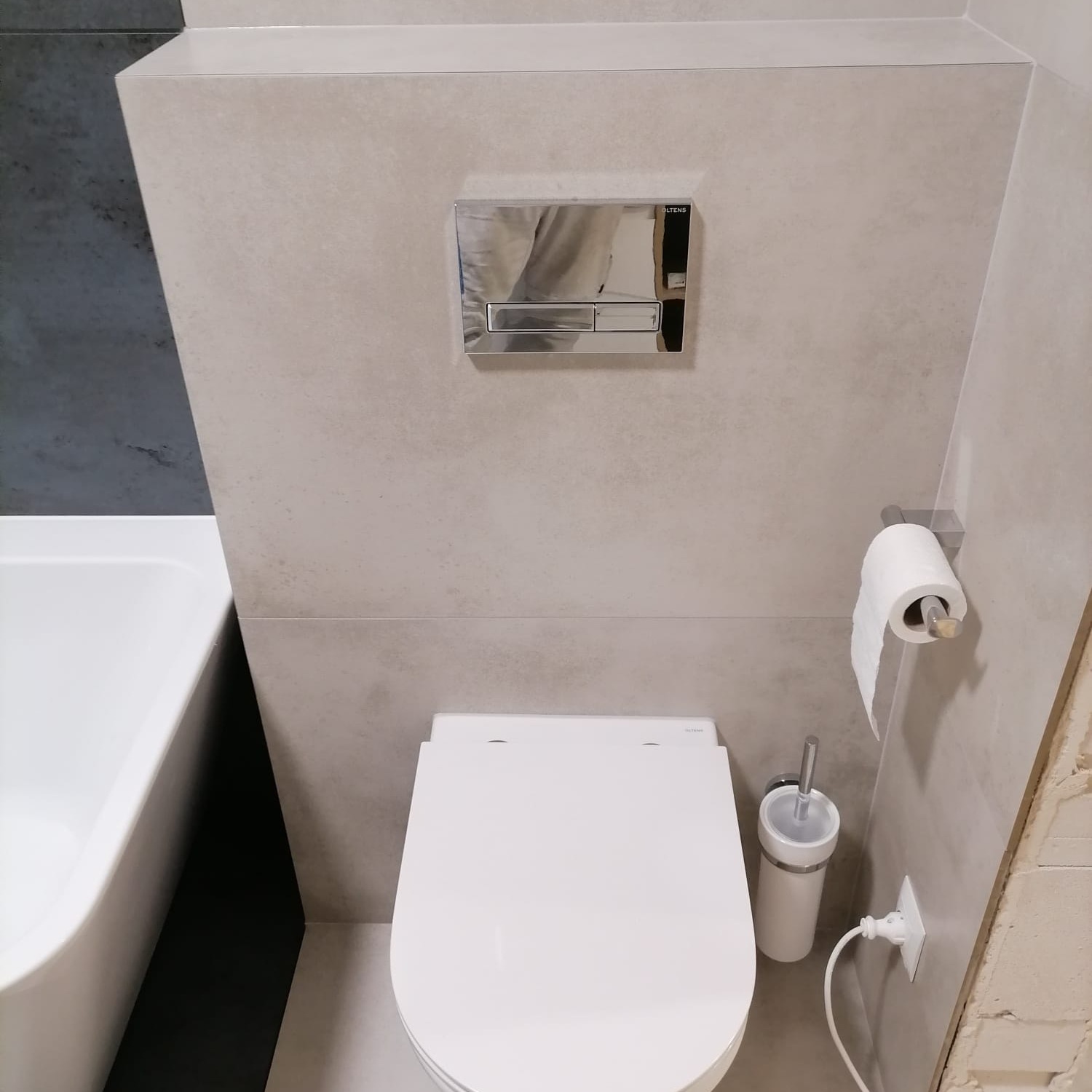 Nowoczesna toaleta podwieszana z chromowanym przyciskiem spłukującym marki Oltens, obok szczotka do WC i uchwyt na papier toaletowy, z widoczną częścią wanny po lewej stronie.