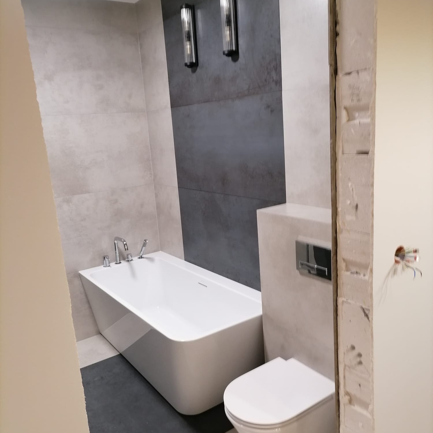 Biała, prostokątna wanna wolnostojąca z chromowaną armaturą, wisząca toaleta z przyciskiem, szare płytki imitujące beton na ścianie, oświetlenie ścienne, widok zza futryny drzwi w trakcie prac...