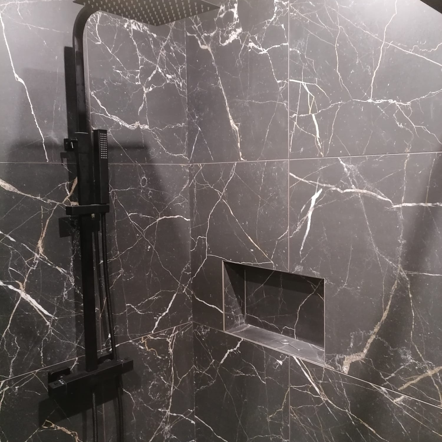 Kabina prysznicowa z czarną armaturą i ścianami wyłożonymi czarnymi płytkami imitującymi marmur z wnęką na kosmetyki.