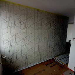 Oktawian Wasiński - Ściana pokryta tapetą w geometryczne wzory, z widoczną taśmą malarską przy suficie, podłoga częściowo zabezpieczona kartonem.
