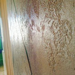 Oktawian Wasiński - Ściana z dekoracyjną, strukturalną farbą w odcieniach brązu i beżu, z widoczną fakturą i efektem połysku, sfotografowana pod kątem, ukazująca fragment ościeżnicy drzwi.