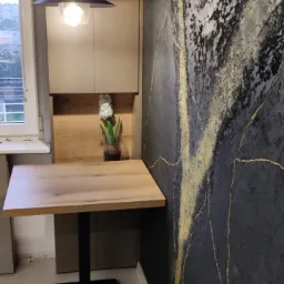 Kompaktowy stół z blatem imitującym drewno, czarna noga, designerska ściana w odcieniach szarości z złotymi akcentami, wisząca lampa w stylu industrialnym, kwiaty w wazonie.