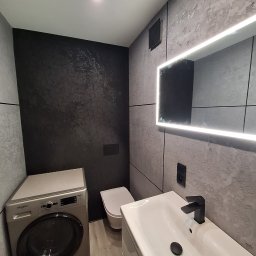 Oktawian Wasiński - Wykończona łazienka w szarościach z podświetlanym lustrem, pralką i umywalką. Na ścianie dekoracyjna, czarna tapeta z geometrycznym wzorem. Nowoczesny design.