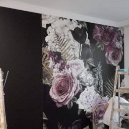 Oktawian Wasiński - Fragment ściany z fototapetą w kwiaty, przylegający do czarnej, gładkiej powierzchni. Ustawiona drabina malarska z narzędziami, osoba w tle podczas pracy.