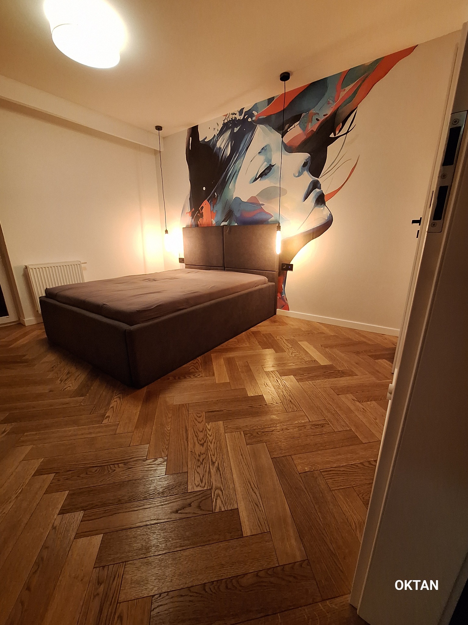 Sypialnia z łóżkiem na parkiecie ułożonym we wzór jodełki, ściana zdobiona abstrakcyjnym malunkiem kobiety, oświetlenie punktowe.