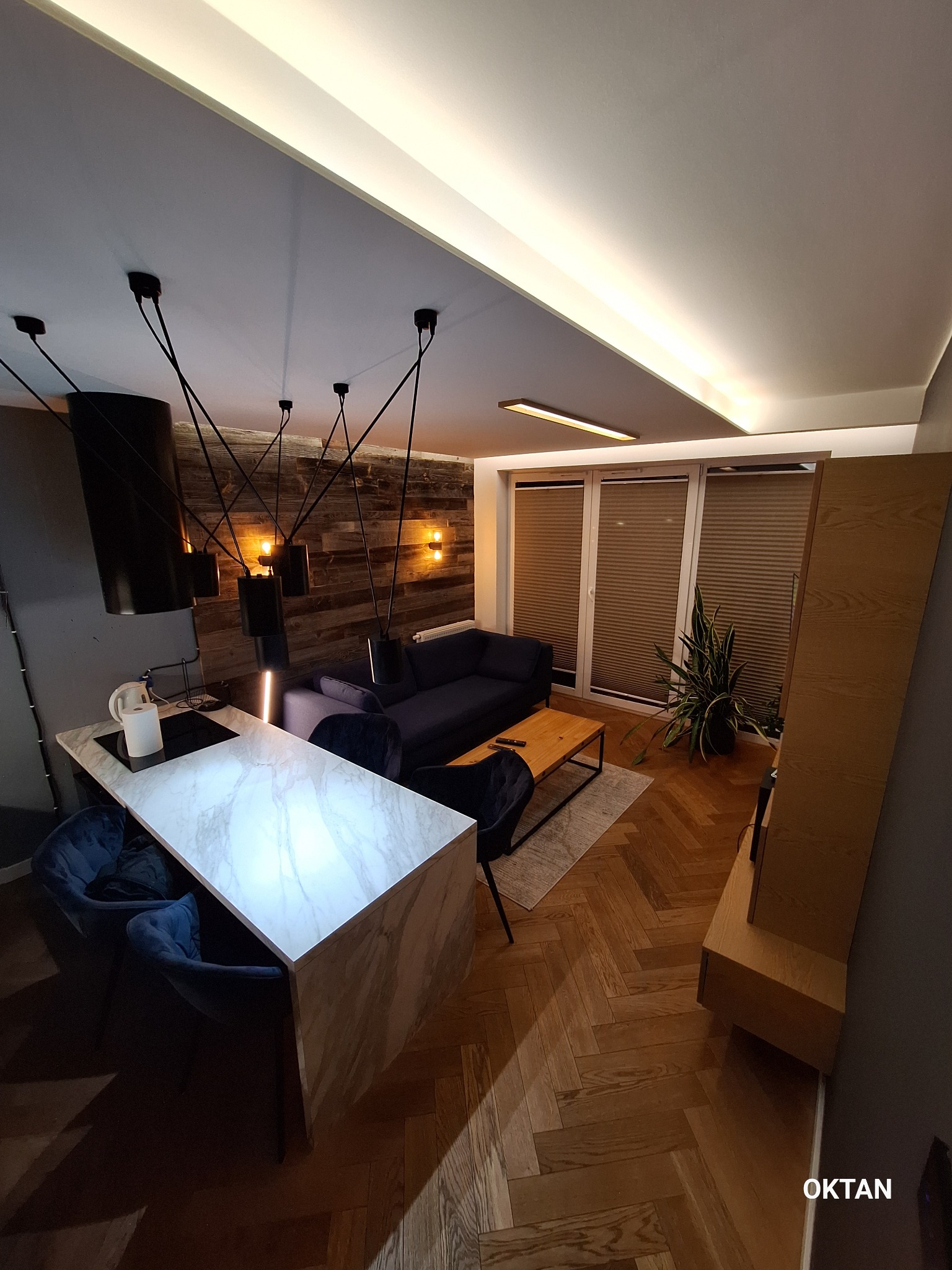 Nowoczesne wnętrze mieszkania z drewnianą ścianą, parkietem w jodełkę, wyspą kuchenną z marmurowym blatem i designerskimi lampami wiszącymi.