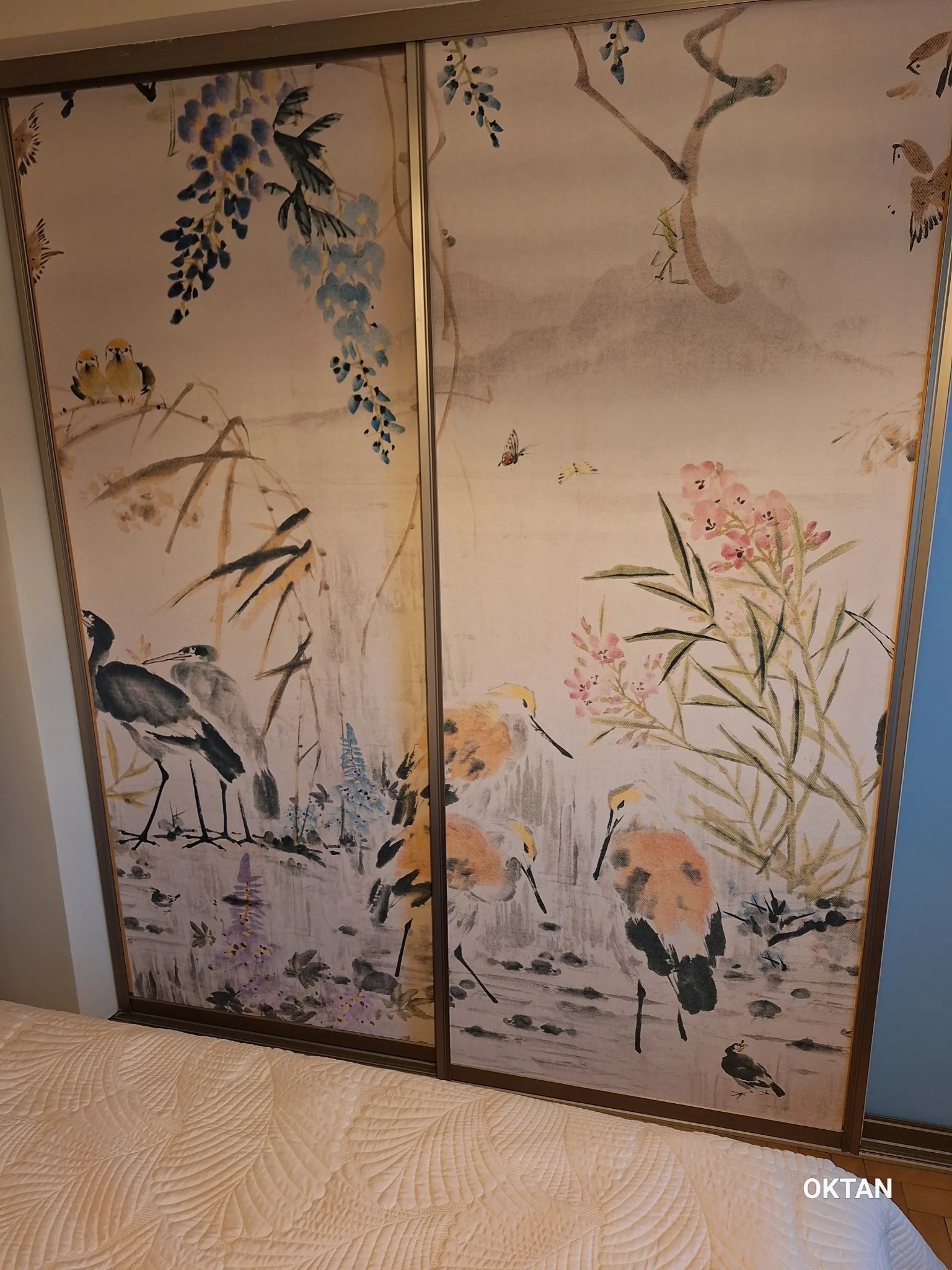 Szafa przesuwna z dekoracyjnym, ręcznie malowanym motywem orientalnym przedstawiającym ptaki, kwiaty i bambus na jasnym tle, widoczna część łóżka z narzutą.