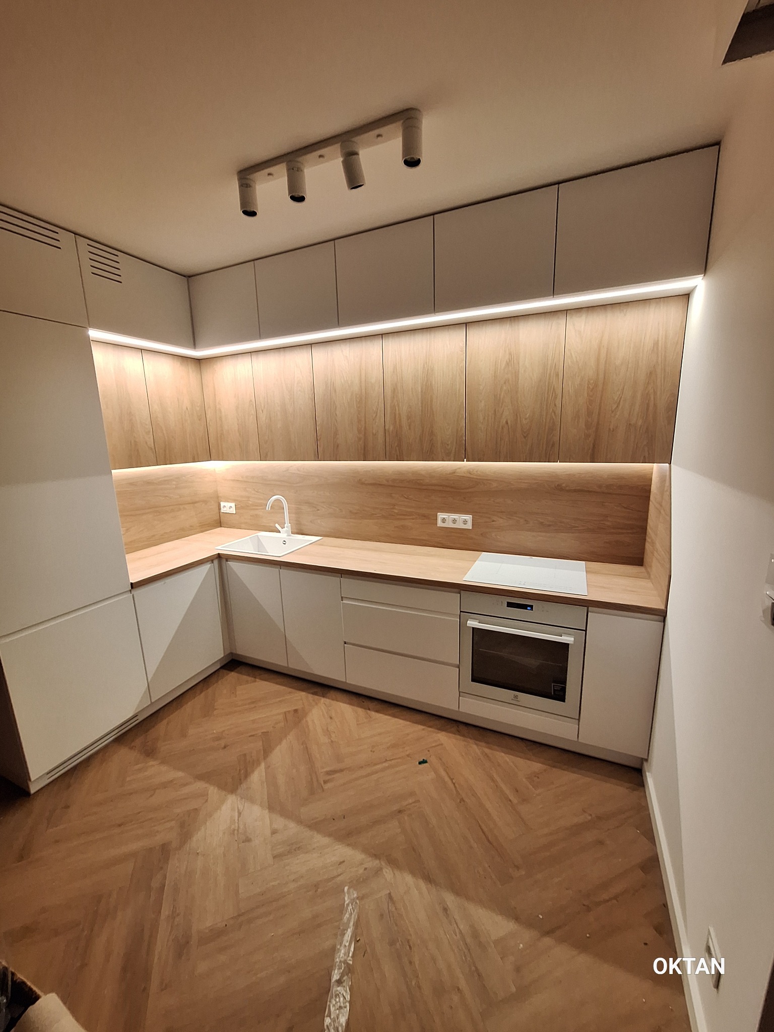 Nowoczesna kuchnia w odcieniach bieli i drewna, z parkietem ułożonym w jodełkę, oświetleniem LED podszafkowym i reflektorami sufitowymi.