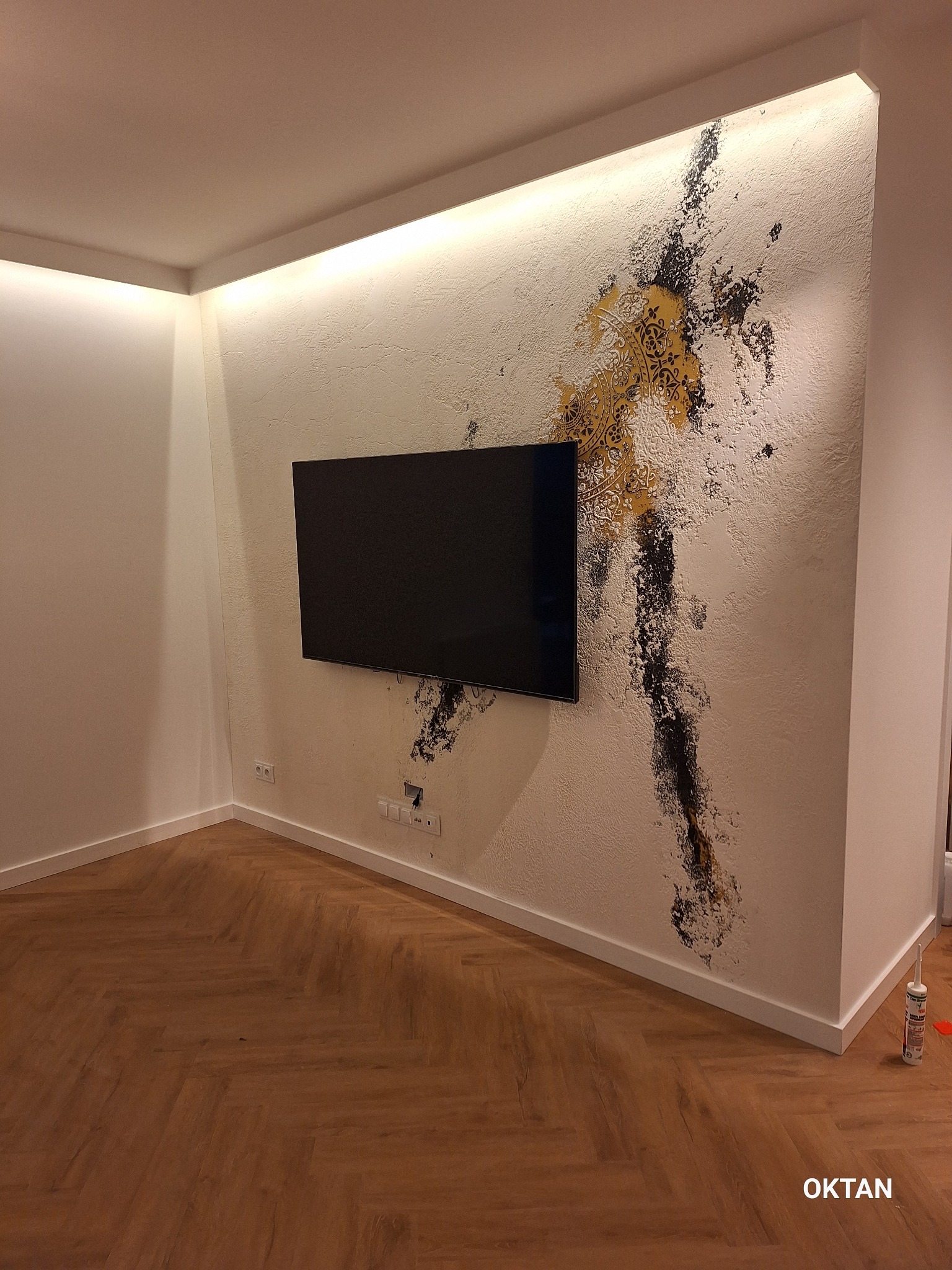 Pokój z wykończoną ścianą z dekoracyjną fakturą i czarno-złotym wzorem, na ścianie zawieszony telewizor, parkiet ułożony w jodełkę, listwa LED podsufitowa.