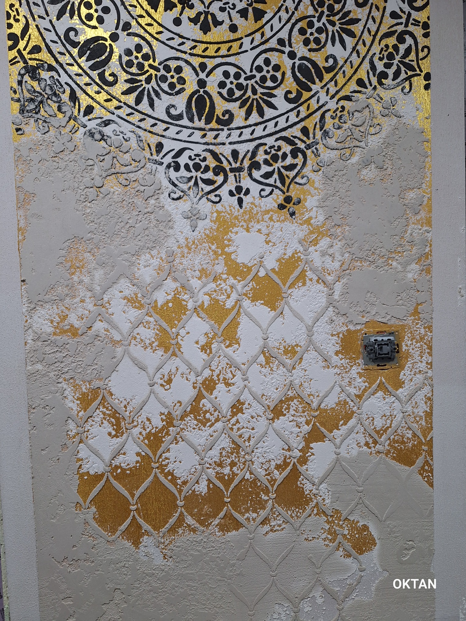 Dekoracyjna ściana z ornamentem mandala w górnej części i wzorem siatki romboidalnej w dolnej, z elementami złocenia i fakturą imitującą nierówny tynk, obok włącznika światła.