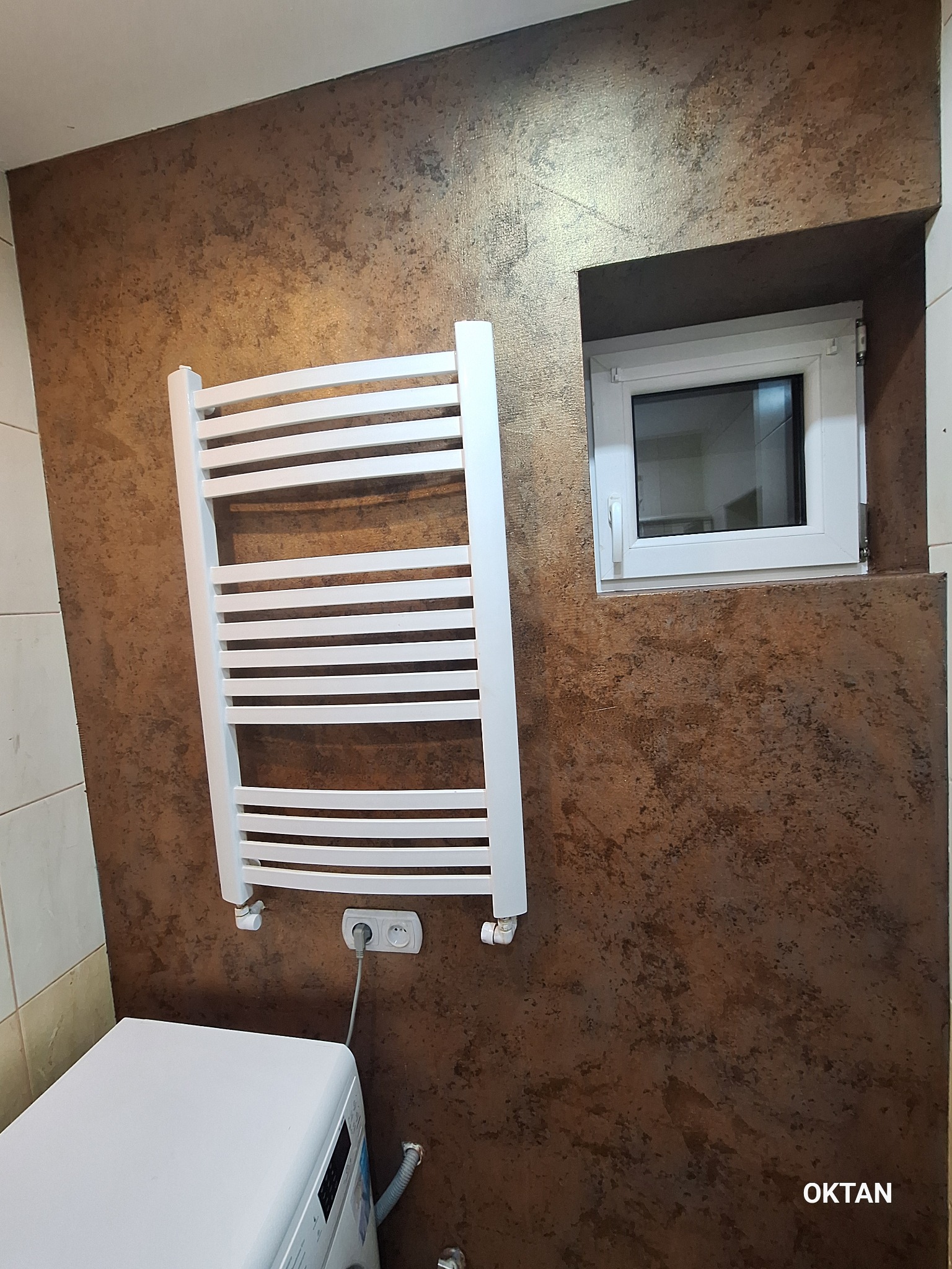 Błyszcząca, brązowa ściana z efektem dekoracyjnym w łazience, z zamontowanym białym grzejnikiem drabinkowym i oknem wnękowym. Widoczny fragment pralki.