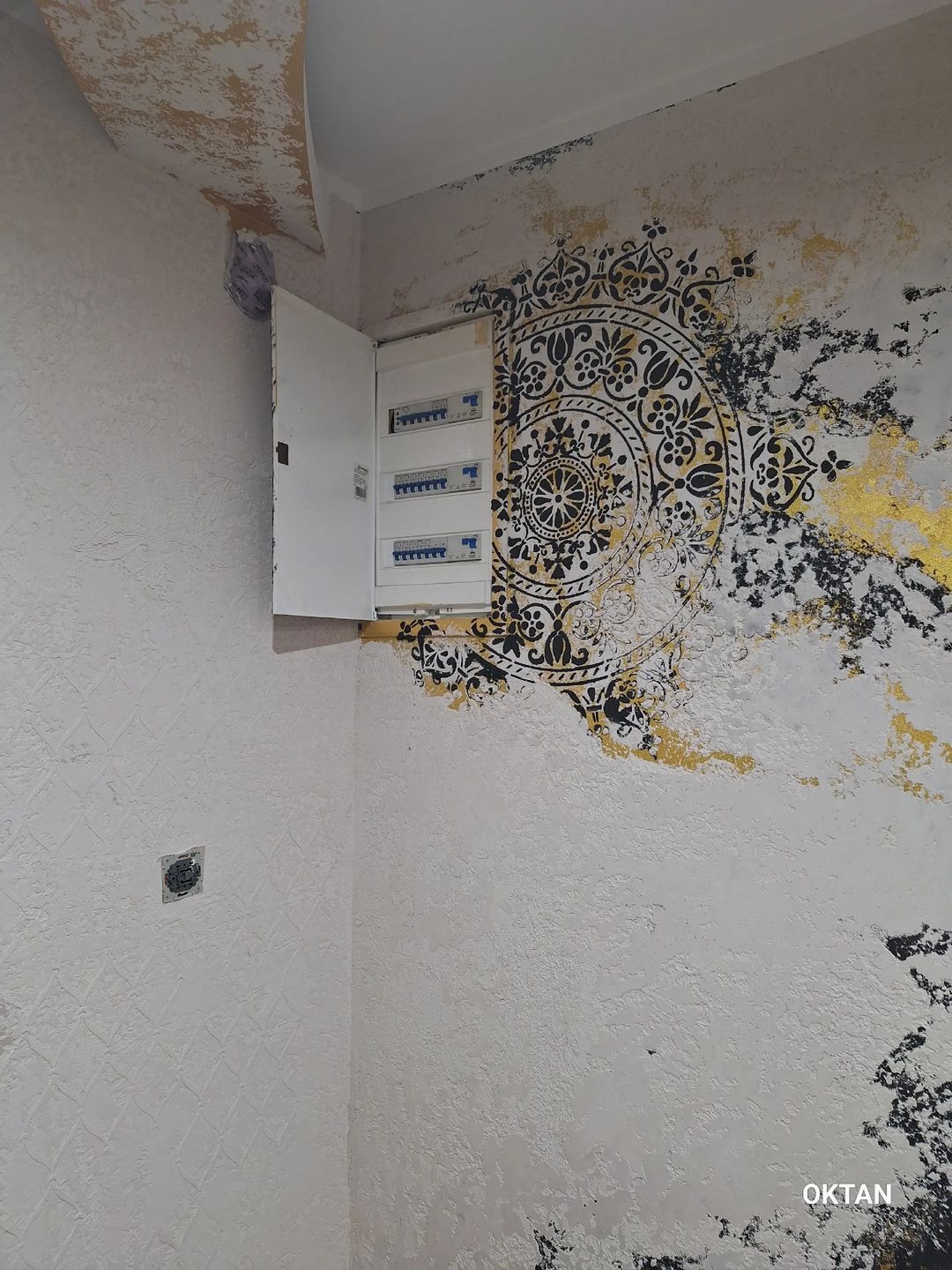 Ściana w rogu pokoju z dekoracyjnym malowaniem: biała struktura, czarny wzór mandali z elementami złota, otwarta skrzynka z bezpiecznikami.